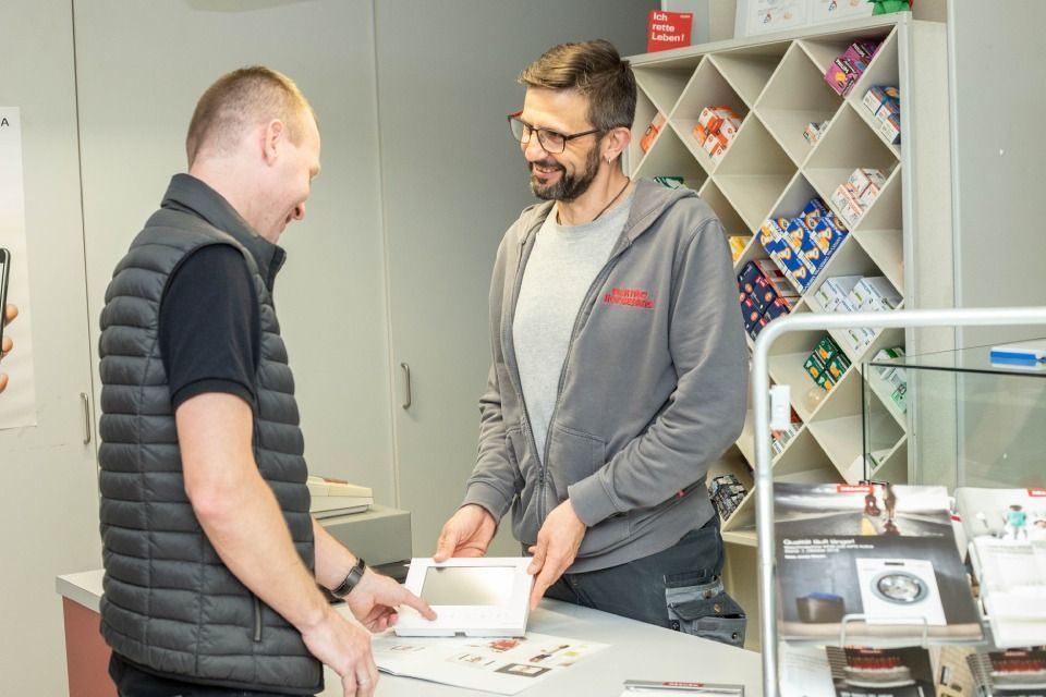 Zwei Männer in einem Geschäft; einer zeigt dem anderen ein Produkt. Im Hintergrund sind Regale mit Artikeln zu sehen.
