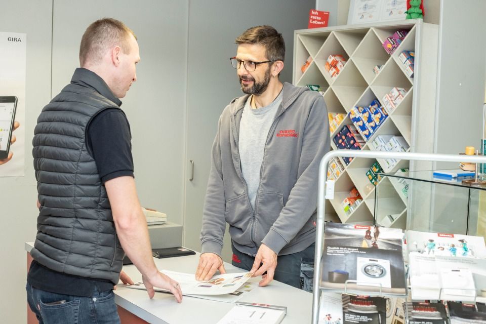 Ein Mann spricht mit einem Verkäufer an einer Theke, möglicherweise in einem Geschäft, hält eine Broschüre in der Hand, im Hintergrund sind Regale voller Artikel.