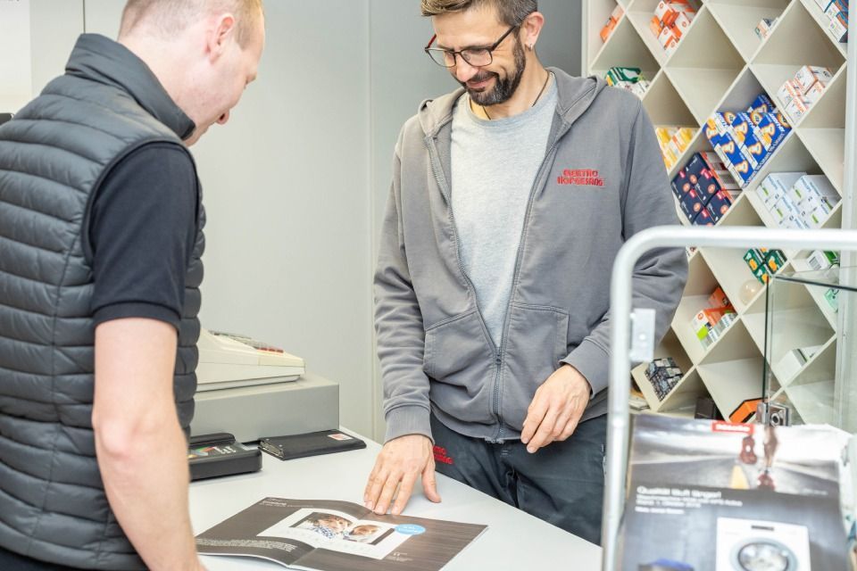 Zwei Männer betrachten ein Dokument auf einem Ladentisch. Einer trägt eine Weste, der andere einen grauen Kapuzenpullover. Im Hintergrund Regale.