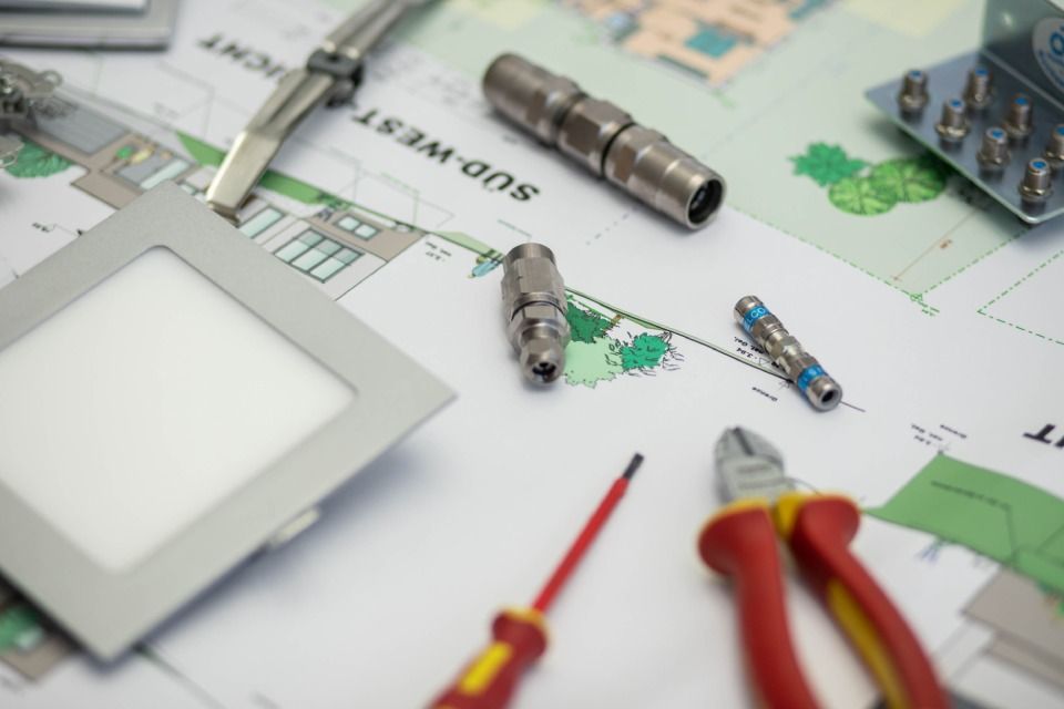 Elektrische Werkzeuge und Komponenten auf einem Bauplan, darunter eine quadratische LED-Leuchte, Stecker und Zangen.