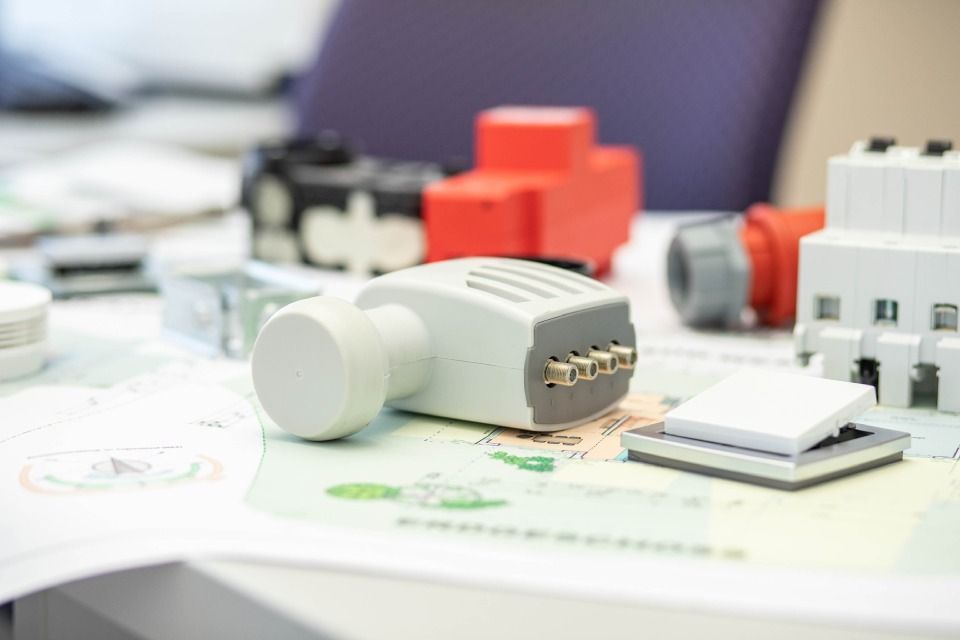 Verschiedene elektrische Komponenten auf einem Tisch, darunter ein hellgrauer Stecker, eine rote Box und weiße Leistungsschalter.