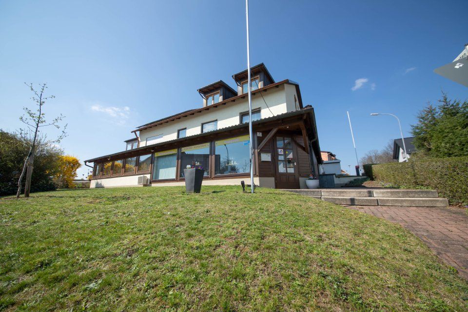 Haus mit braunen Zierleisten und großen Fenstern auf einem grasbewachsenen Hügel unter blauem Himmel.
