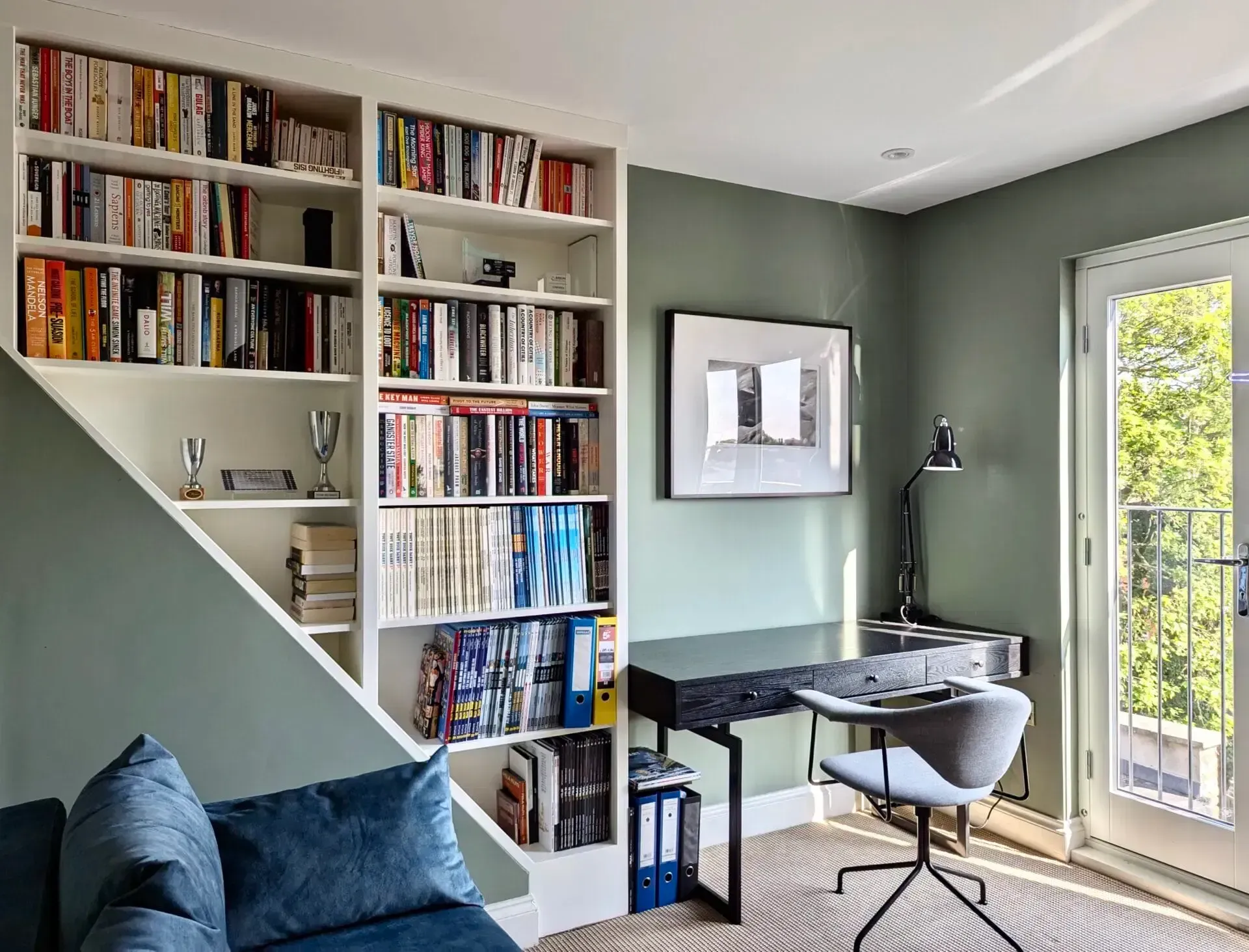 Modern home office with built-in bookshelf, desk, chair, and large window beside a blue sofa
