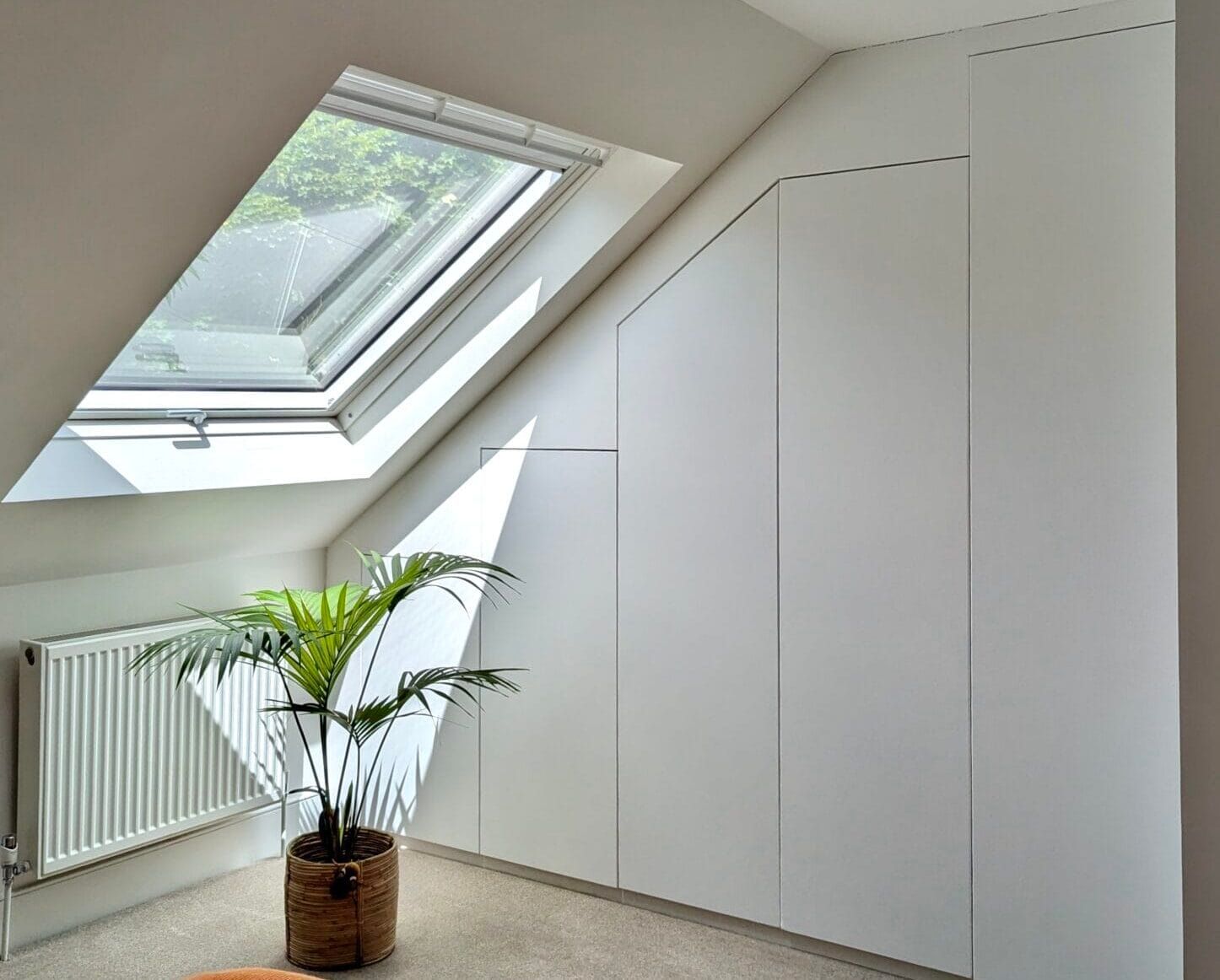 Bright attic room with skylight, white built-in cabinets, radiator, and potted palm on carpeted floor
