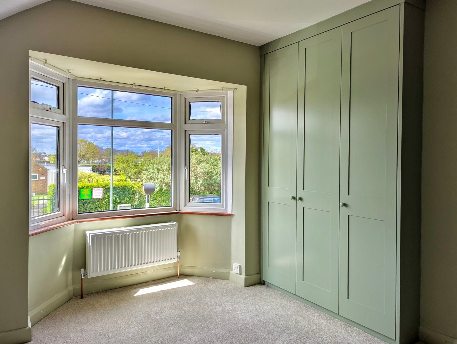 Bright empty bedroom with bay window, white radiator, and built-in pale green wardrobes