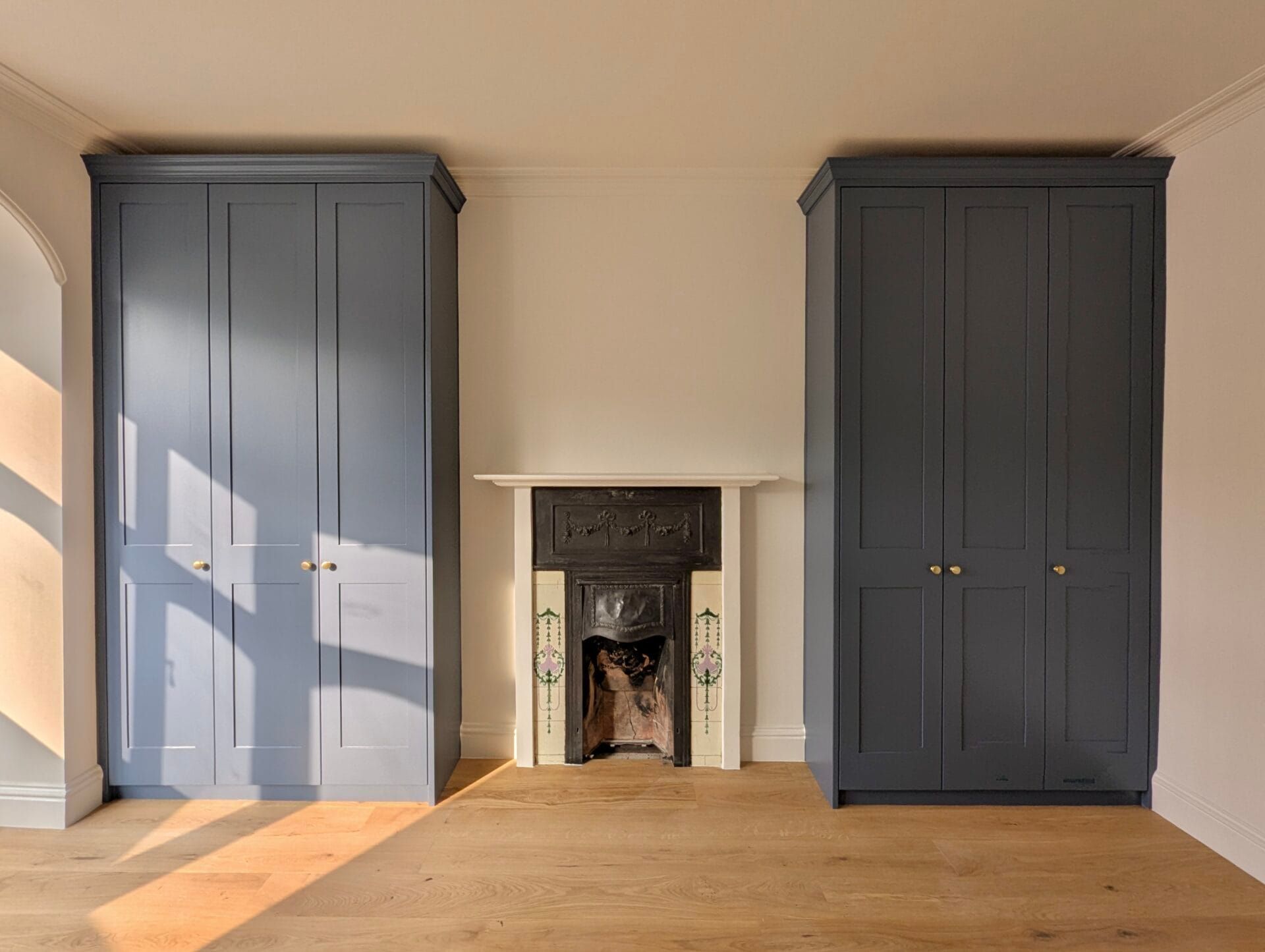 Room with two dark built-in wardrobes and a small fireplace in the center, lit by sunlight on the wooden floor