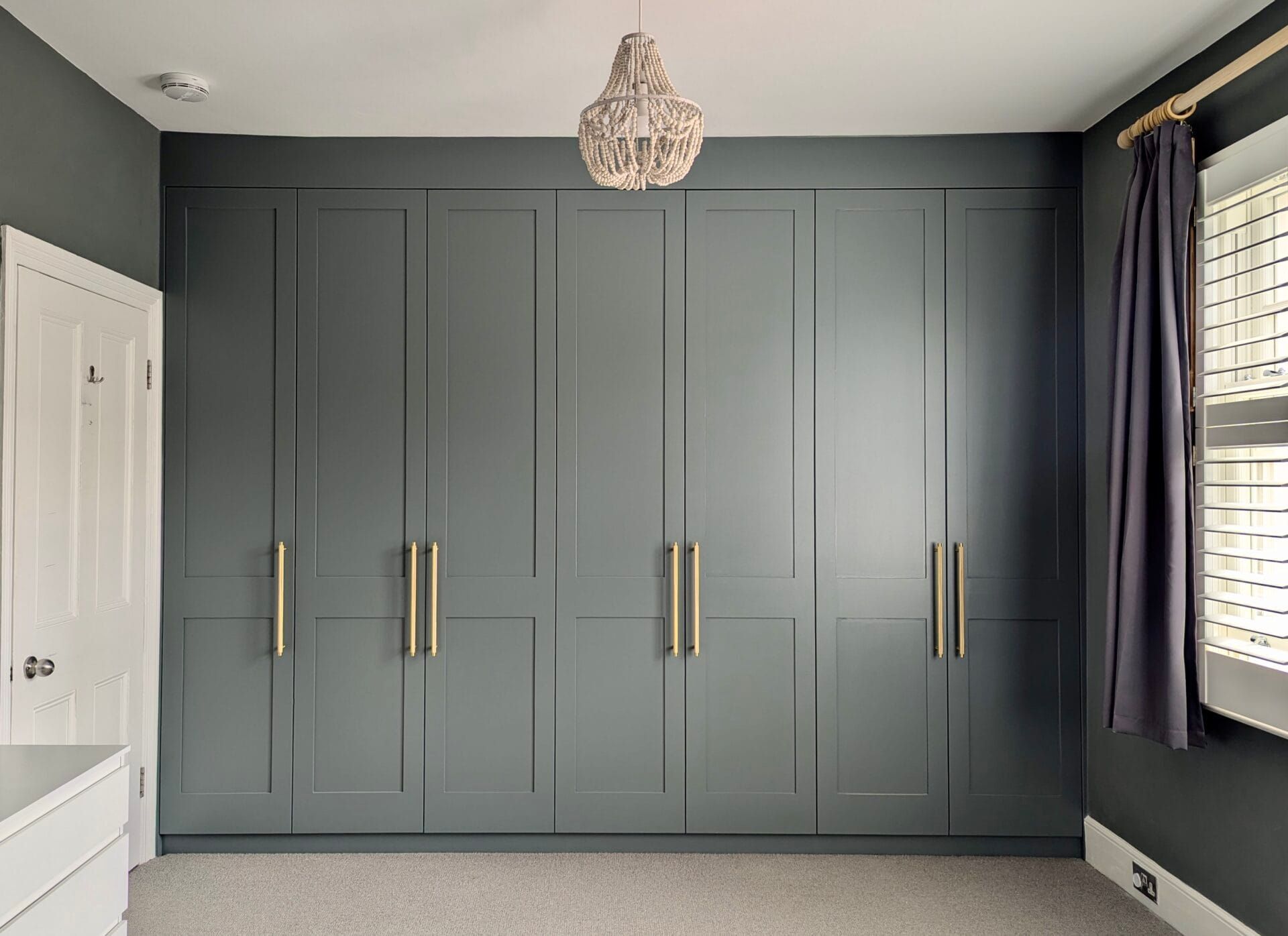 Gray built-in wardrobe with brass handles in a bedroom, beside a window with dark curtains