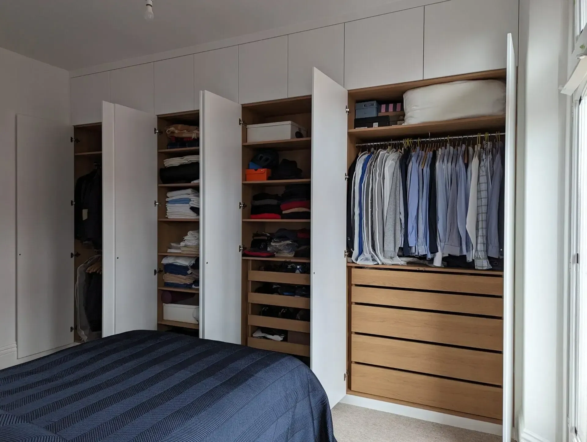 Bedroom with open closets and drawers, showing neatly hung clothes and folded items beside a bed.