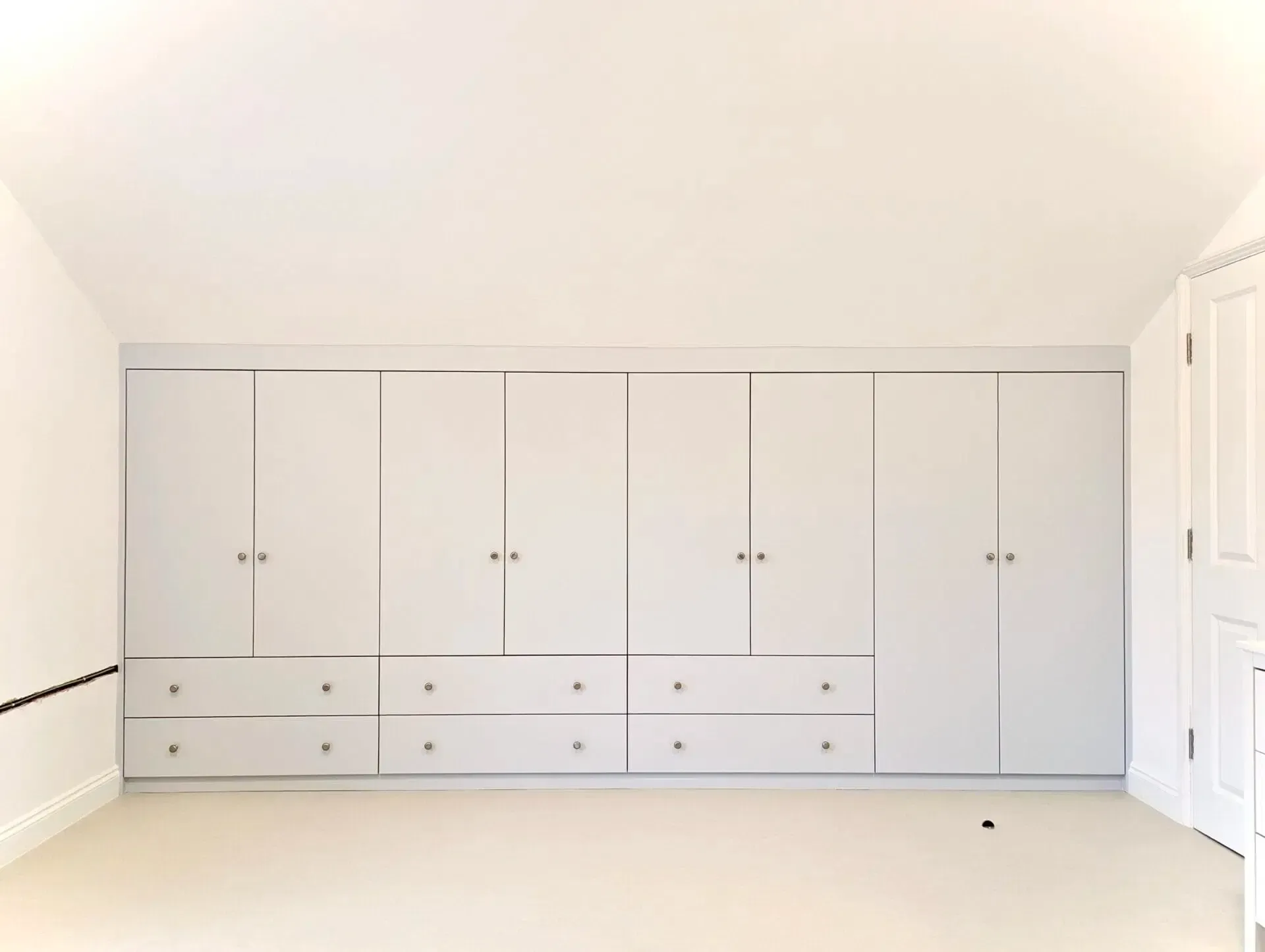 White built-in bedroom wardrobes with drawers and cabinet doors along one wall in a bright room.