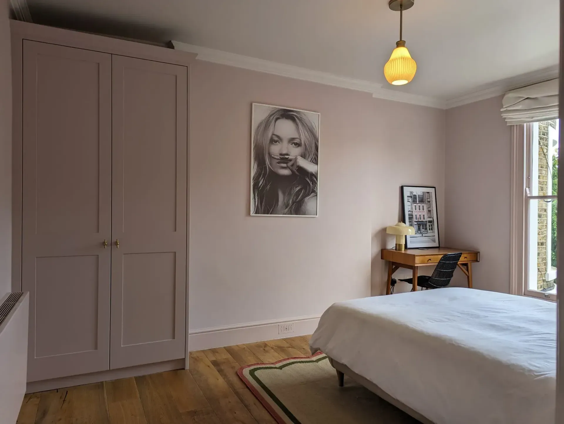 Bright bedroom with a bed, wardrobe, portrait on the wall, and a desk by the window.