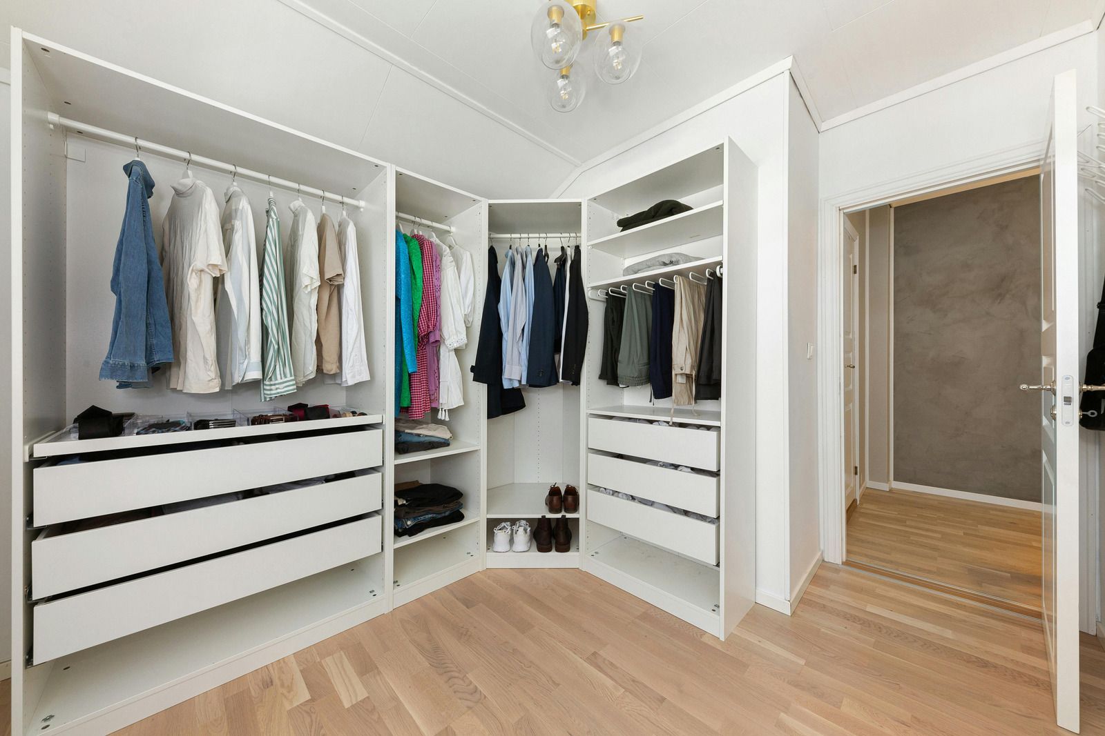 Bright walk-in closet with white shelving, hanging clothes, drawers, and light wood floors.