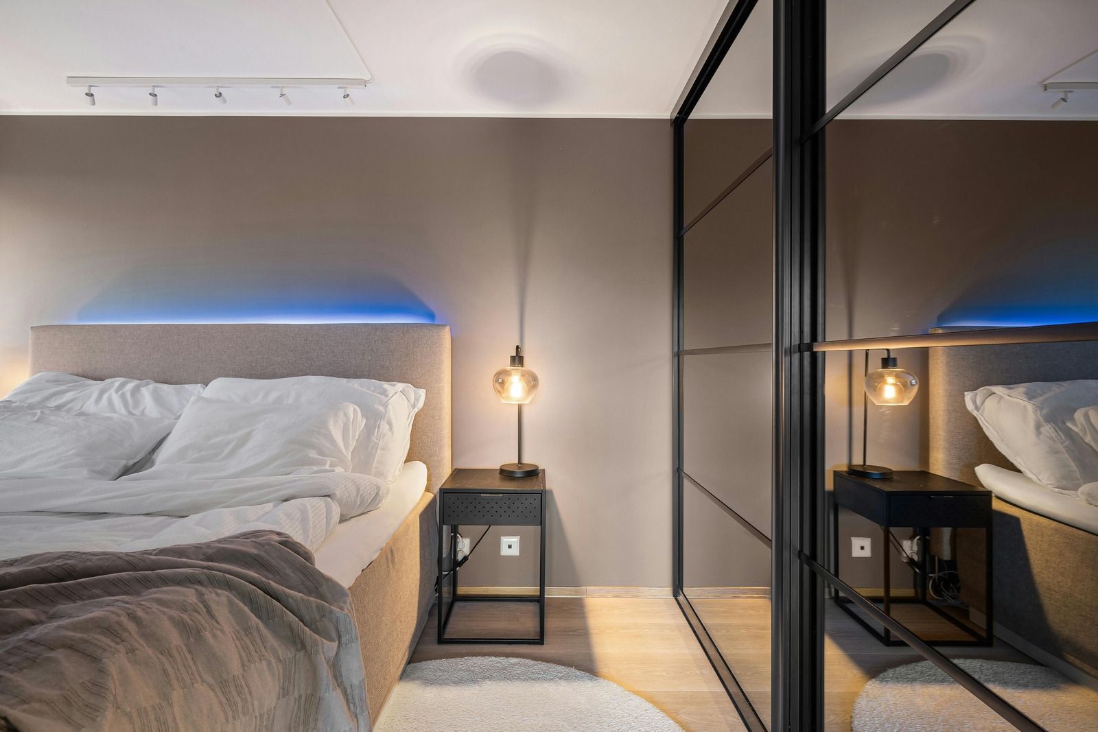 Modern bedroom with unmade bed, bedside lamp, and mirrored wardrobe with blue accent lighting