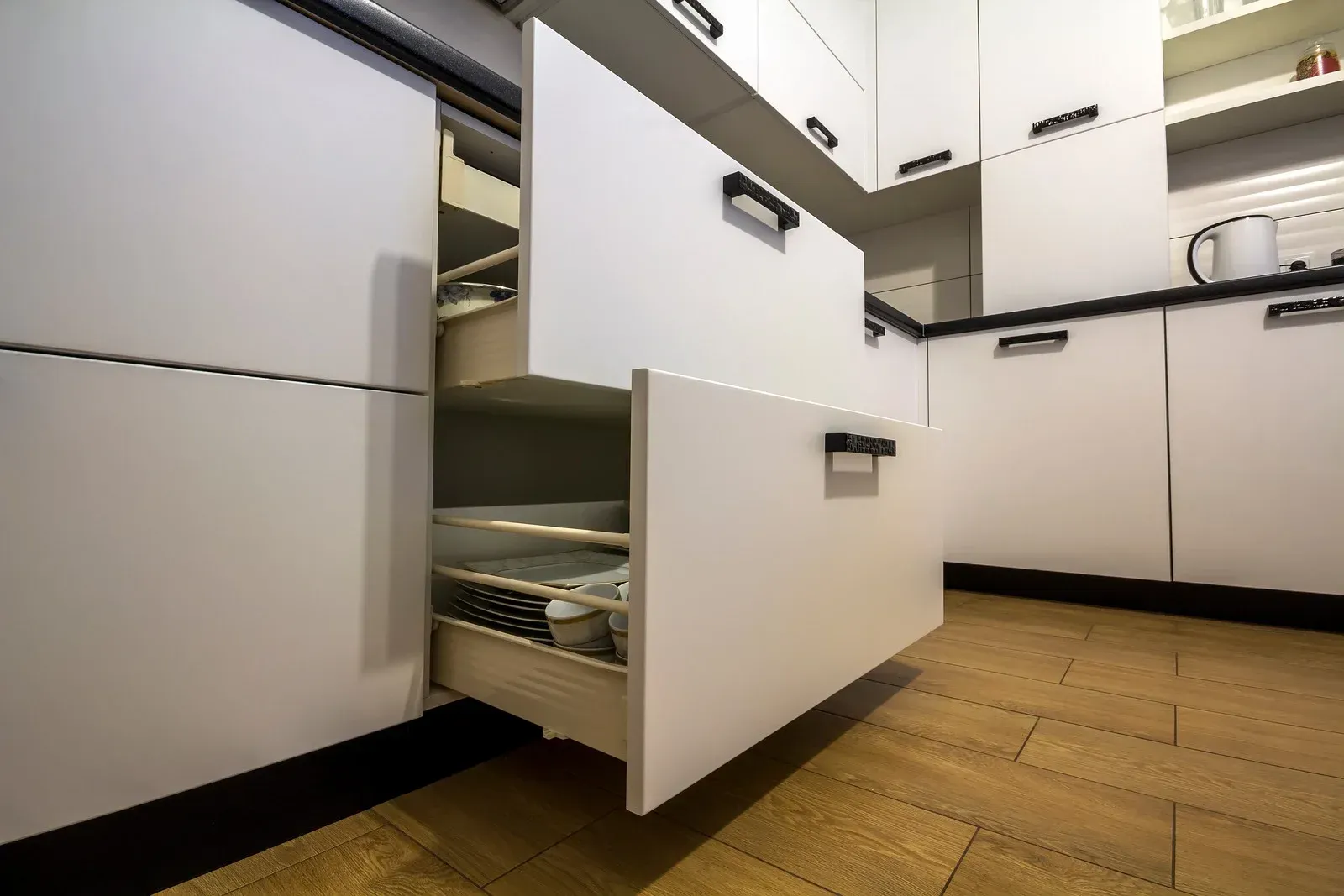 Open kitchen drawer with utensils beside white cabinets and wooden floor