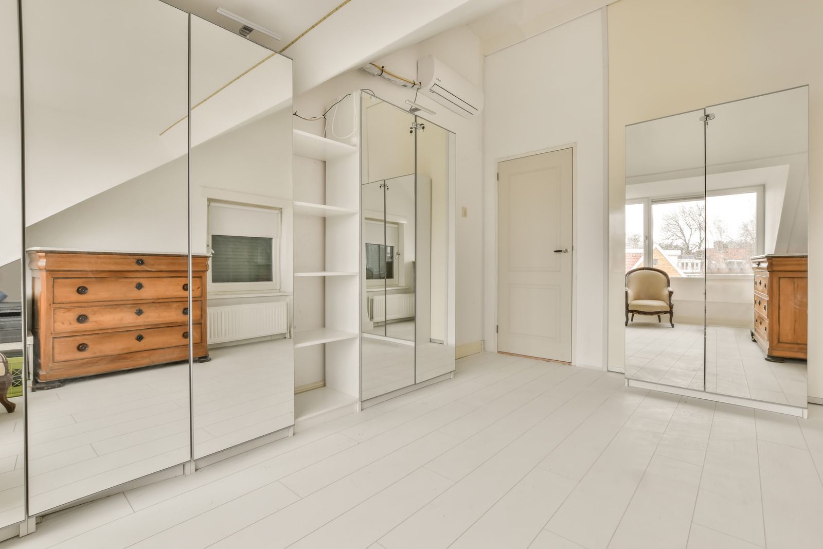 Bright white bedroom with mirrored closet doors, wooden dresser, and open doorway to a sunlit room