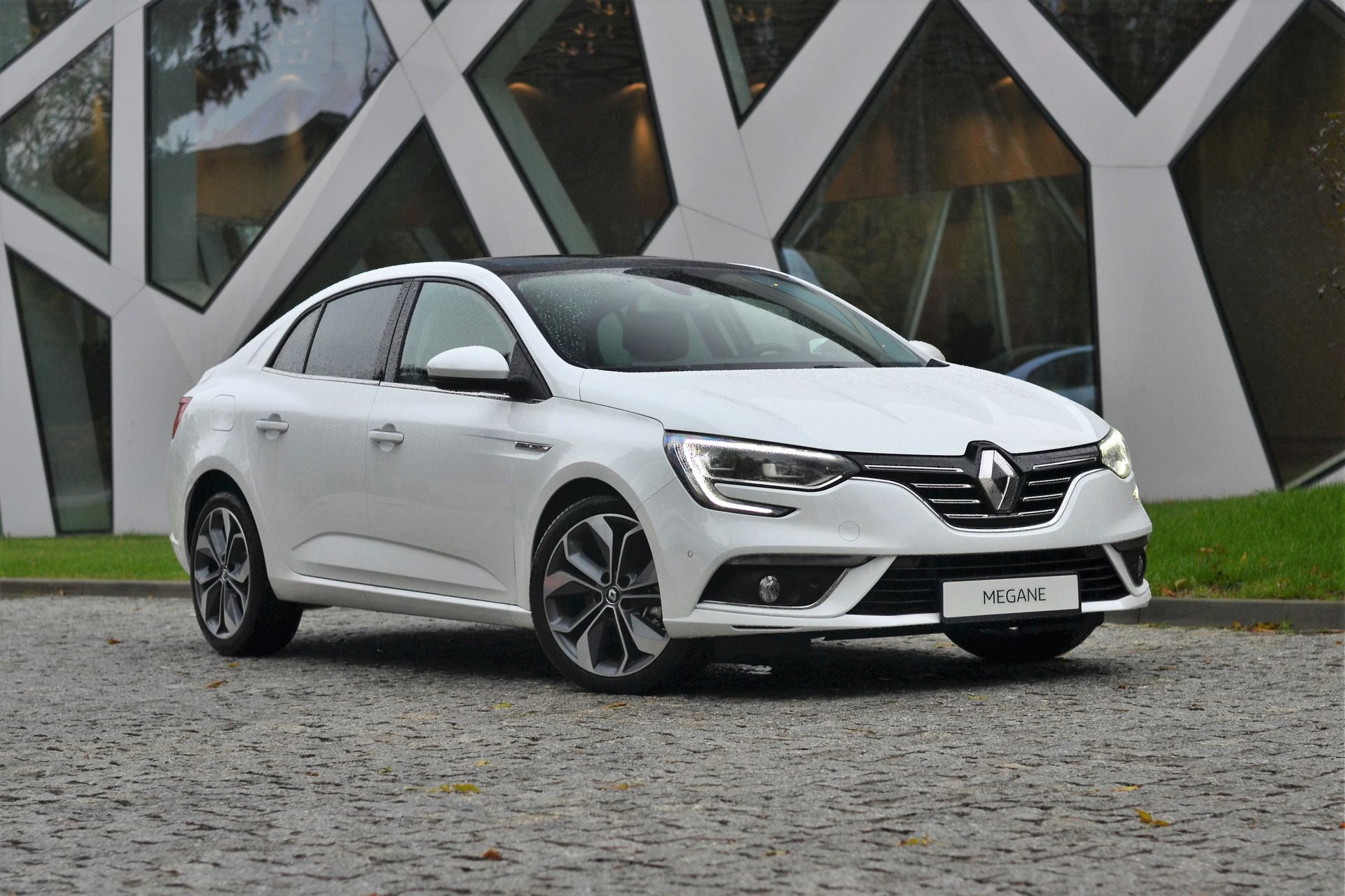 Renault Megane sedán blanco estacionado frente a un edificio moderno con diseño geométrico.