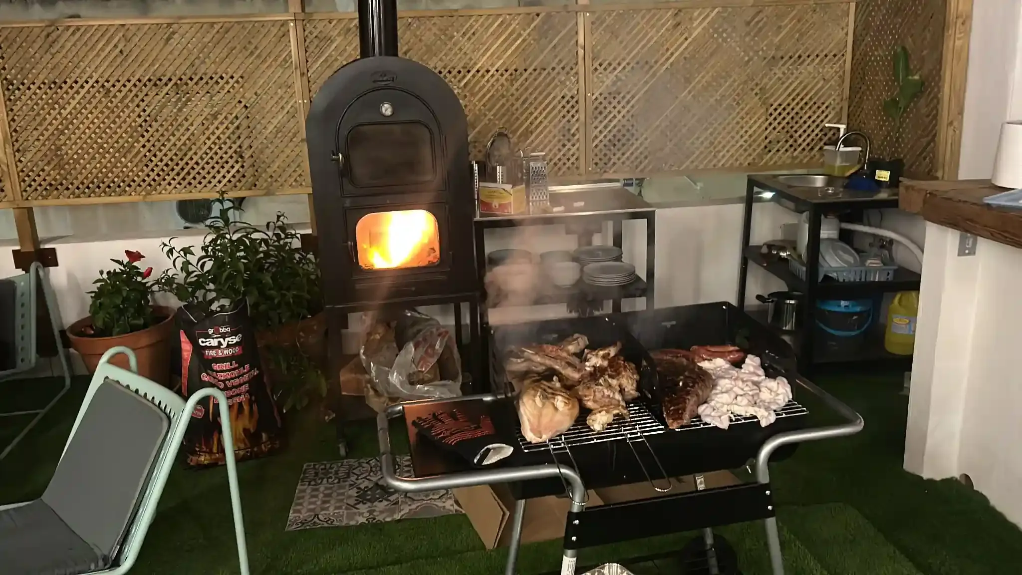 Parrilla con cocción de carnes variadas; una estufa de leña con llama abierta cercana en un espacio al aire libre.
