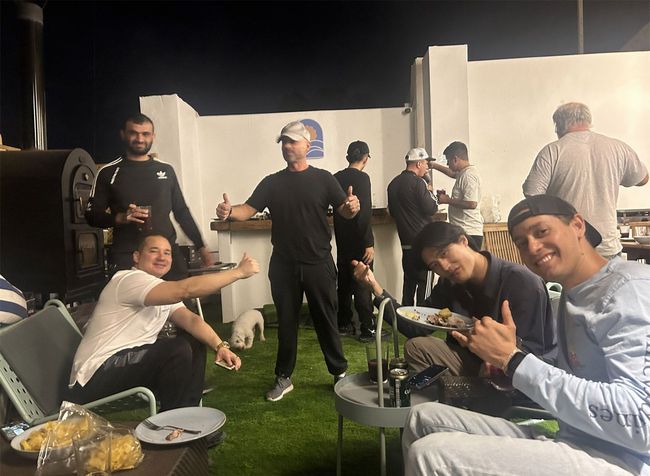 Grupo de personas al aire libre por la noche, pulgares hacia arriba, algunos comiendo en una mesa, césped artificial.