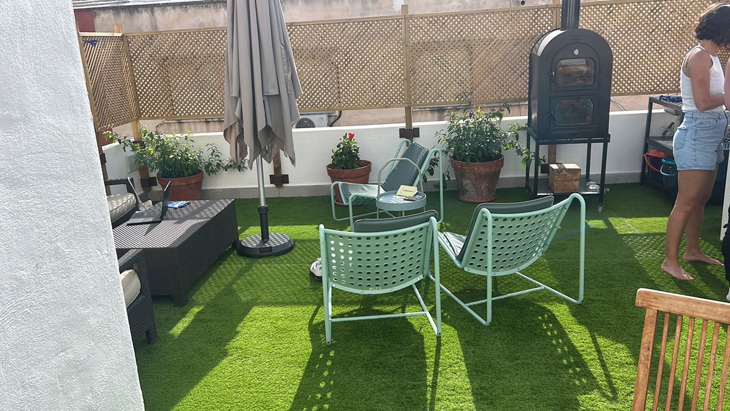 Patio en la azotea con césped verde, asientos, sombrilla y una persona cerca de un horno al aire libre.