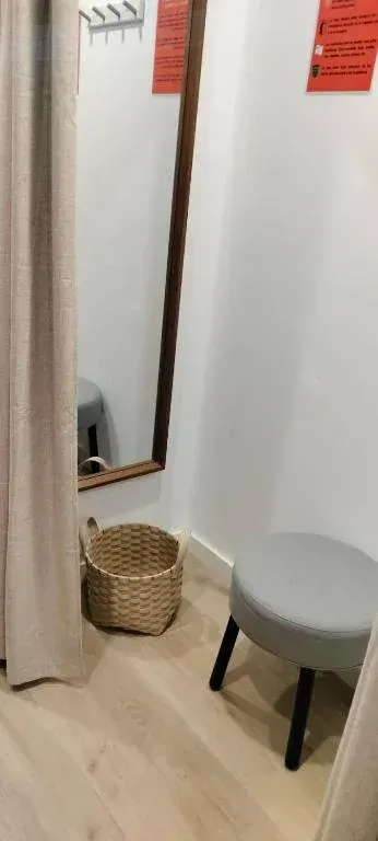 Probador con espejo, cesta tejida y taburete. Cortina beige y paredes blancas.
