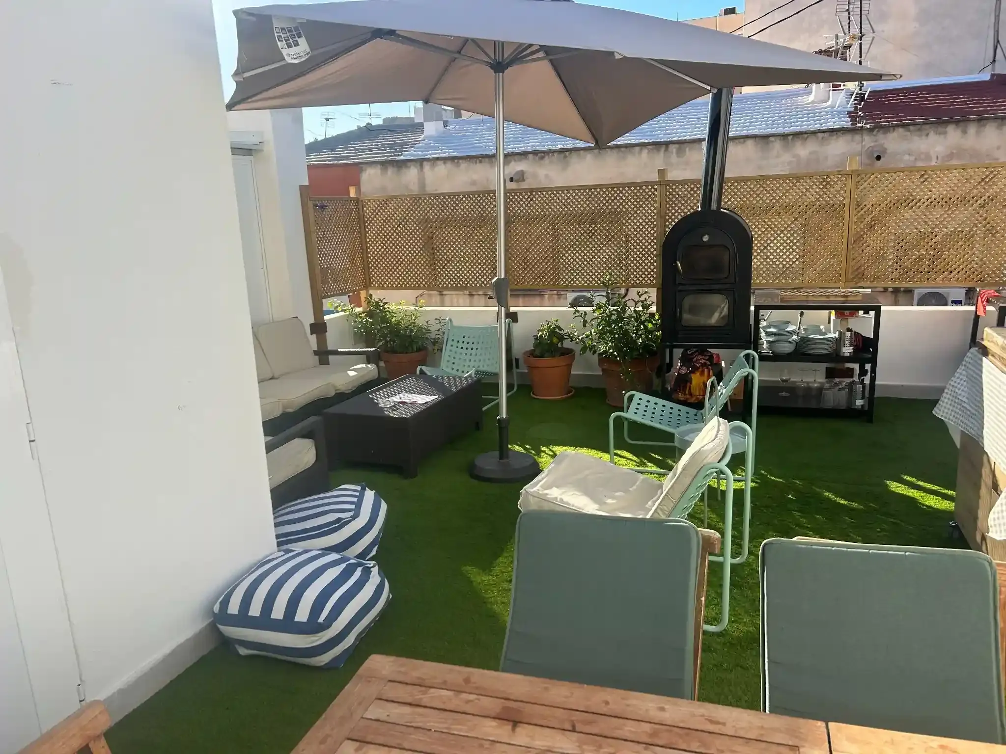 Patio en la azotea con asientos, sombrilla y barbacoa. Césped artificial verde, tonos neutros y luz natural.