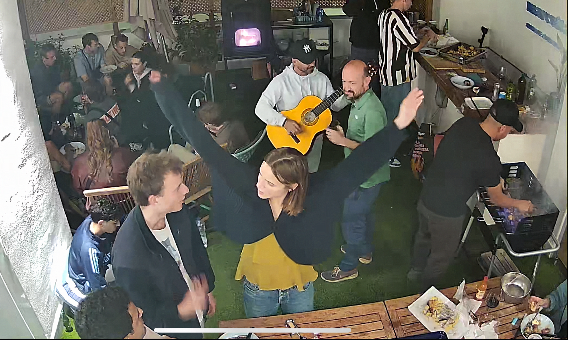 Gente celebrando en una reunión. Una mujer con los brazos en alto, un guitarrista y otros cerca de mesas con comida y equipo. Interior.