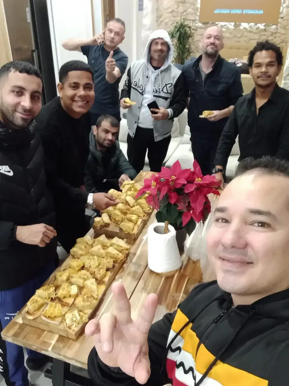 Grupo de hombres sentados a una mesa con comida, algunos con platos. Ambiente relajado y sonriente.
