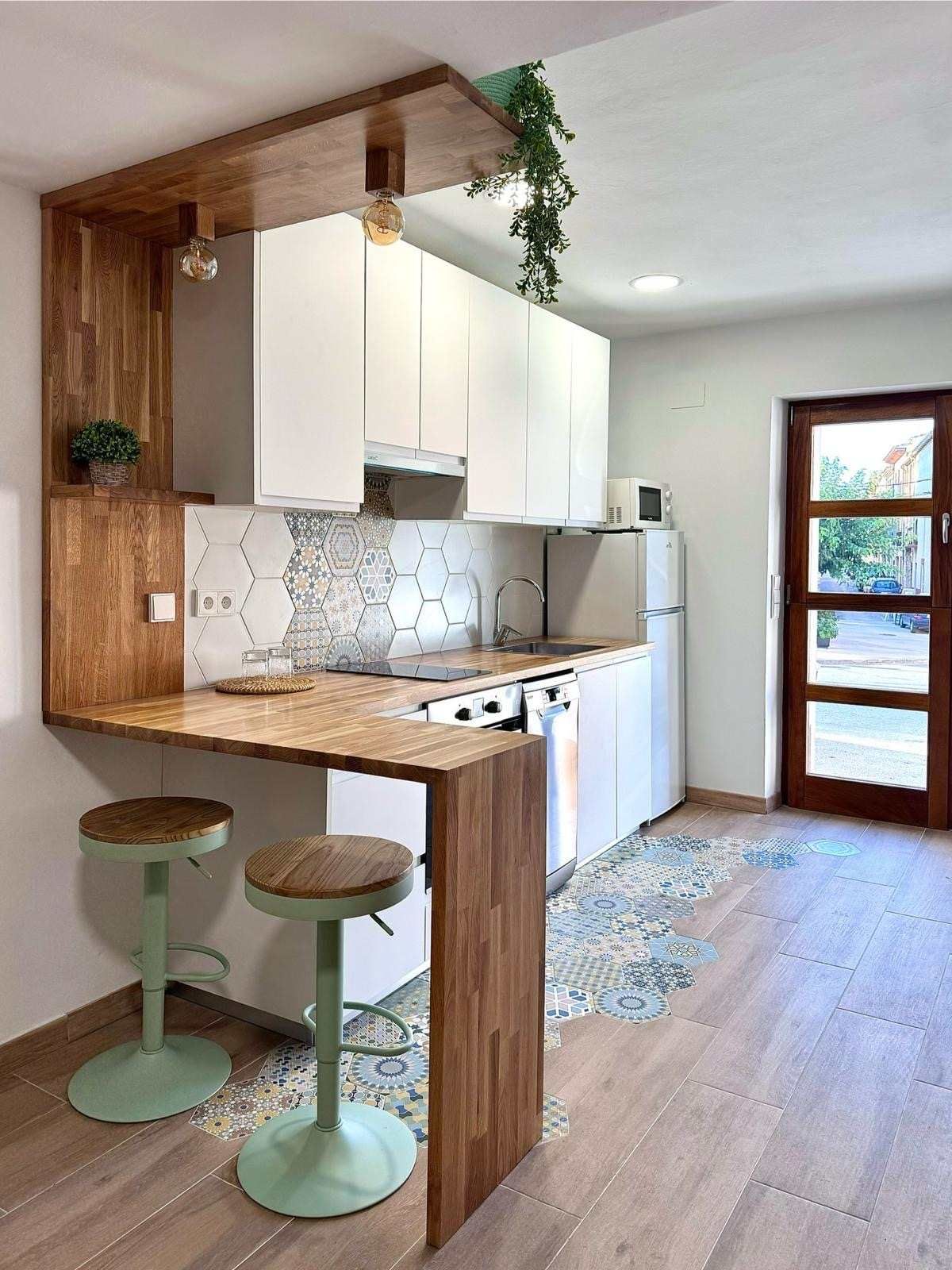 Cuina moderna amb barra d'esmorzar de fusta, armaris blancs, tamborets verd menta i una porta a l'exterior.