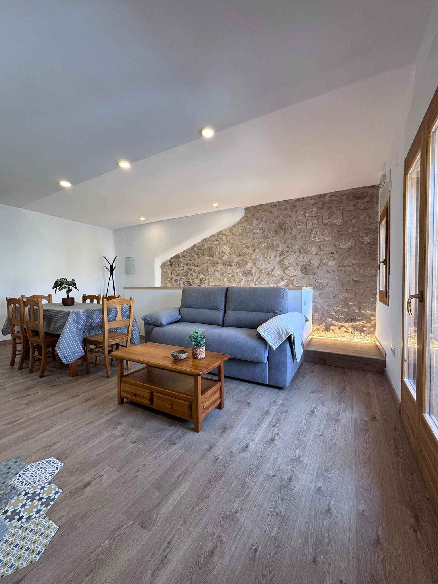 Sala d'estar de planta oberta amb paret de pedra, taula de menjador, sofà gris, taula de centre de fusta i finestres.