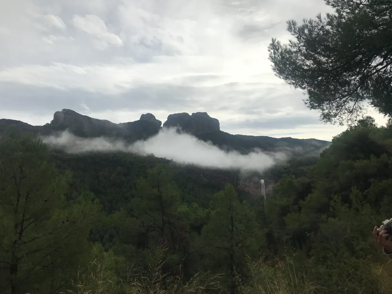 Muntanyes parcialment tapades pels núvols, envoltades d'arbres verds sota un cel ennuvolat.