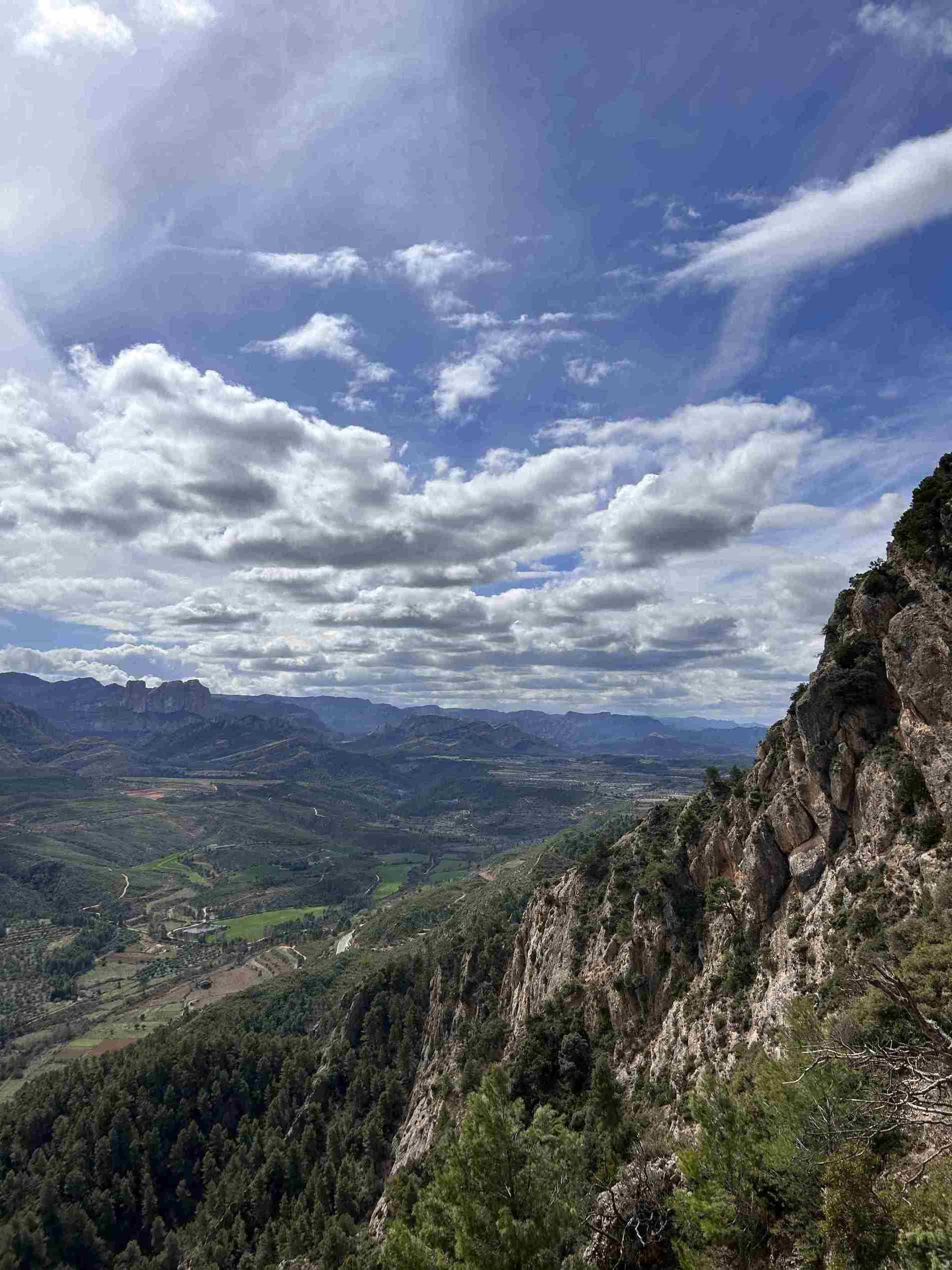 Vista d'una vall i muntanyes, cel blau amb núvols, amb un penya-segat rocós en primer pla.