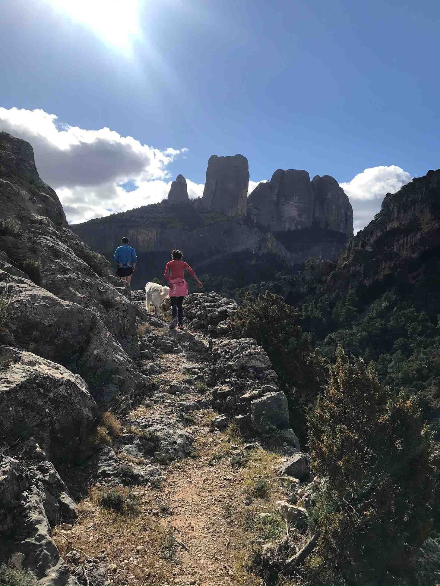 Gent fent senderisme per un camí rocós amb un gos, grans formacions rocoses al fons, dia assolellat.