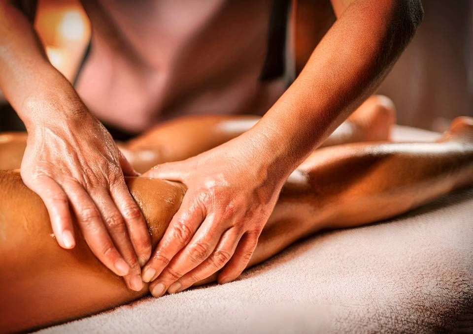 Des mains massent une jambe sur un lit blanc, lumière tamisée.