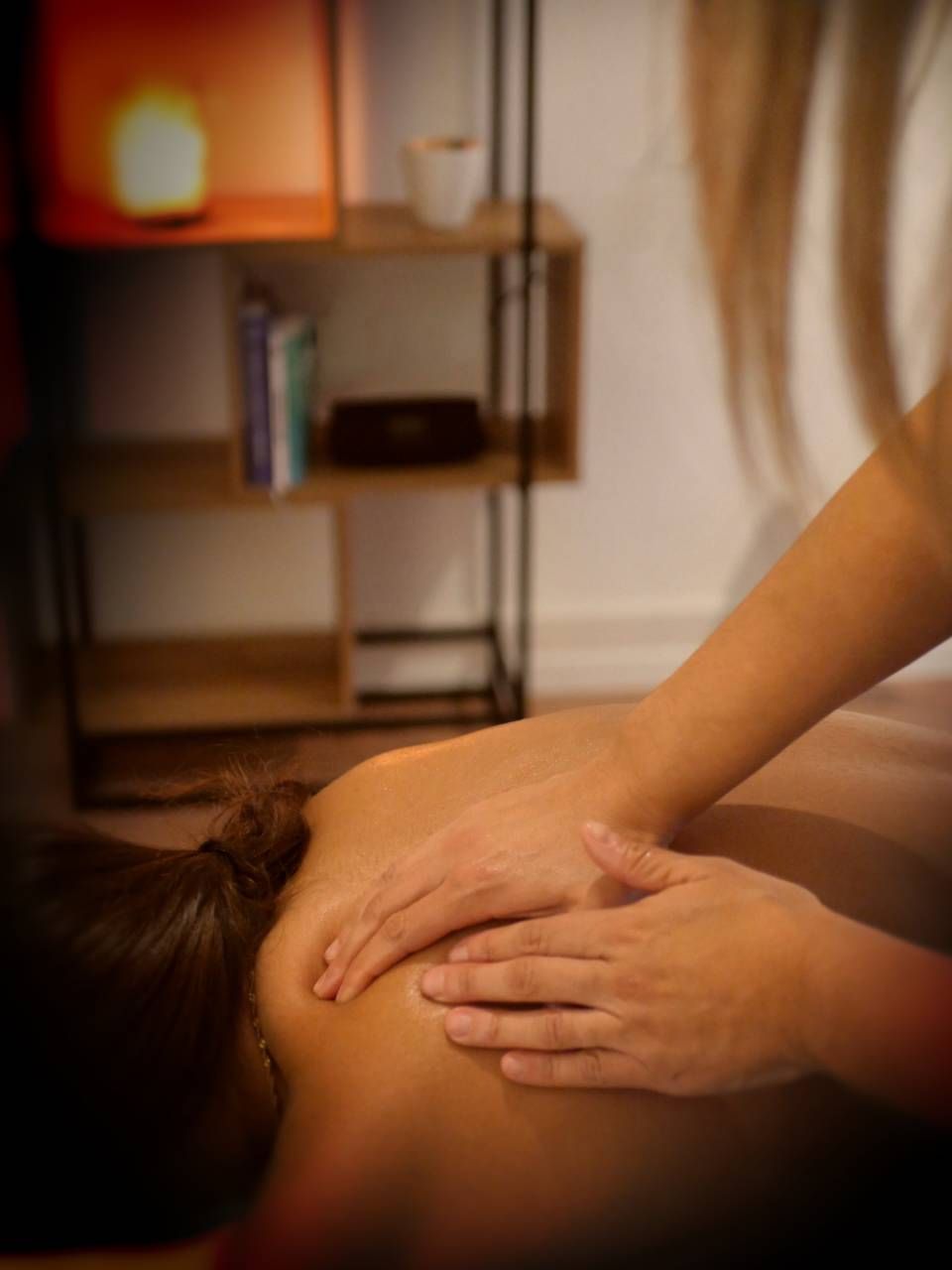 Des mains massent le dos d'une personne. À l'arrière-plan, une étagère avec des livres et une lampe.