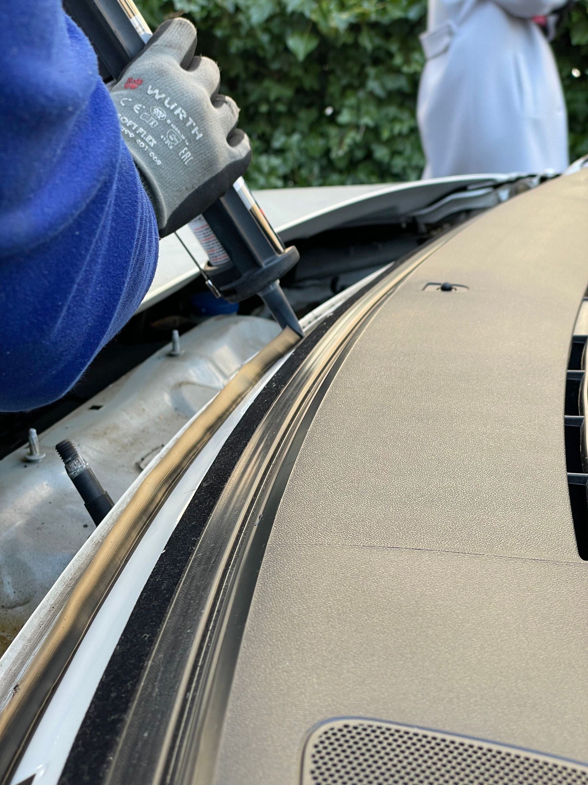 Main gantée utilisant un outil pour étanchéifier un joint sur le toit beige d’une voiture, près du pare-brise