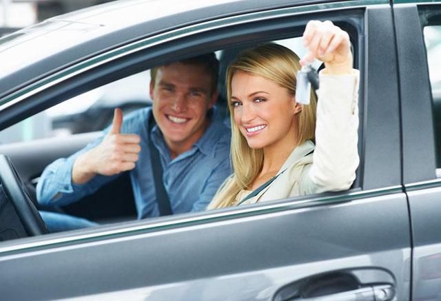Deux personnes sourient et font un pouce levé à l’intérieur d’une voiture grise