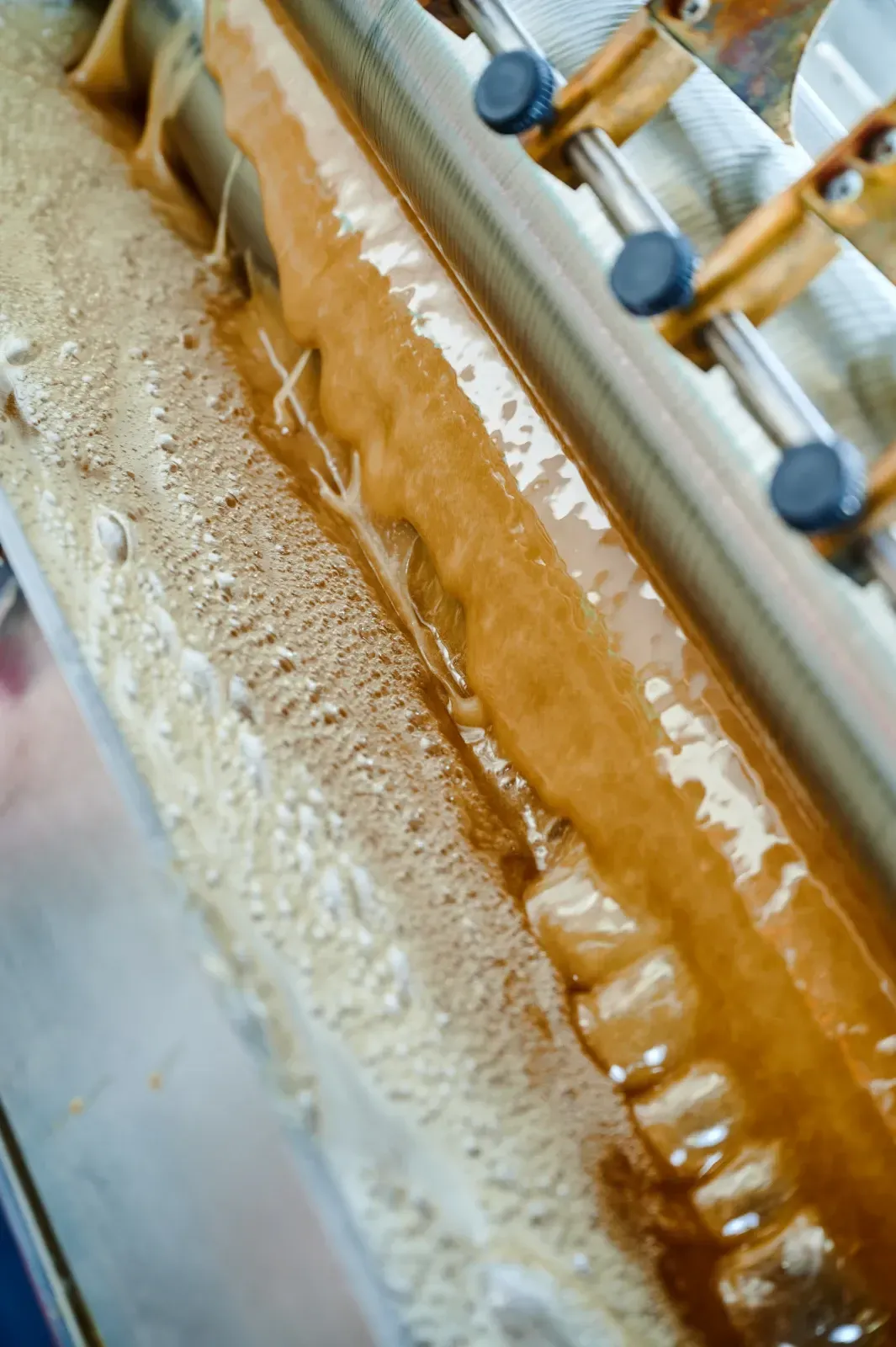Nahaufnahme einer Industriemaschine, die eine beige, schaumige Flüssigkeit auf einem Metallförderband transportiert.