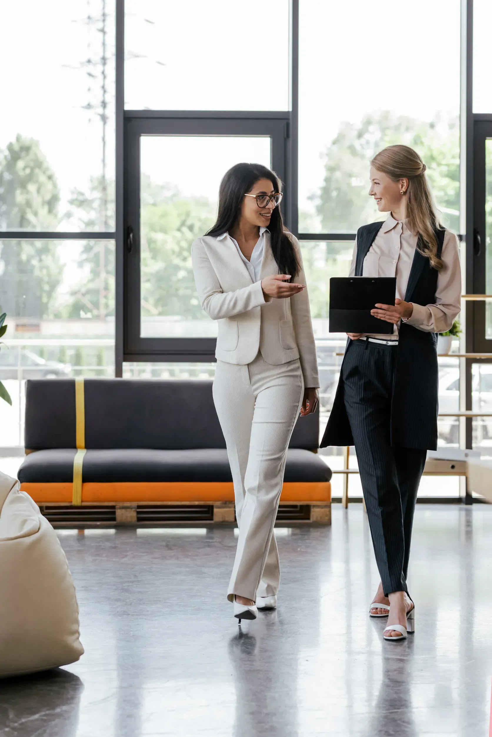 Dos mujeres con atuendos de oficina caminan y conversan; una señala