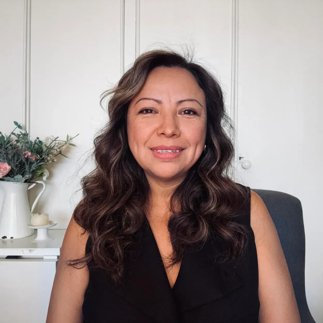 Una mujer con cabello castaño largo y ondulado sonríe a la cámara, sentada frente a una pared blanca.