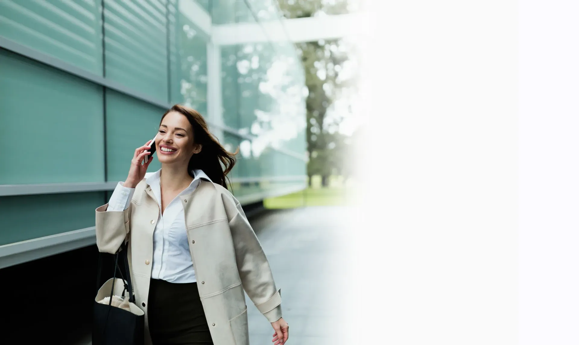 Mujer sonriendo y hablando por teléfono celular, caminando cerca de un edificio moderno con un maletín.