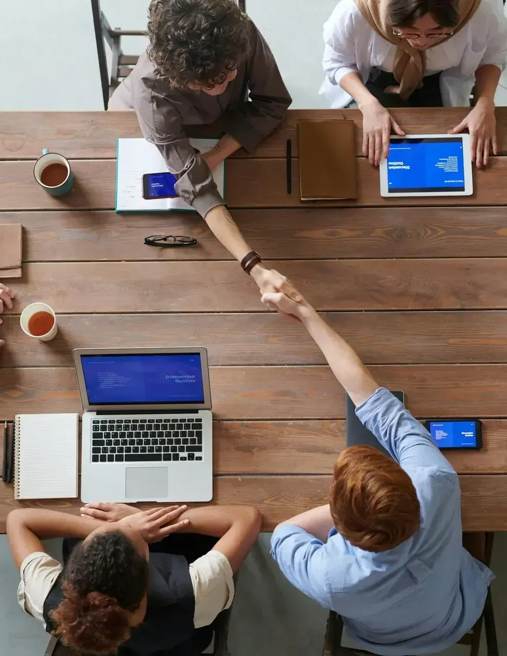 Personas se dan la mano en una mesa de madera con computadoras portátiles, tabletas y tazas de café.