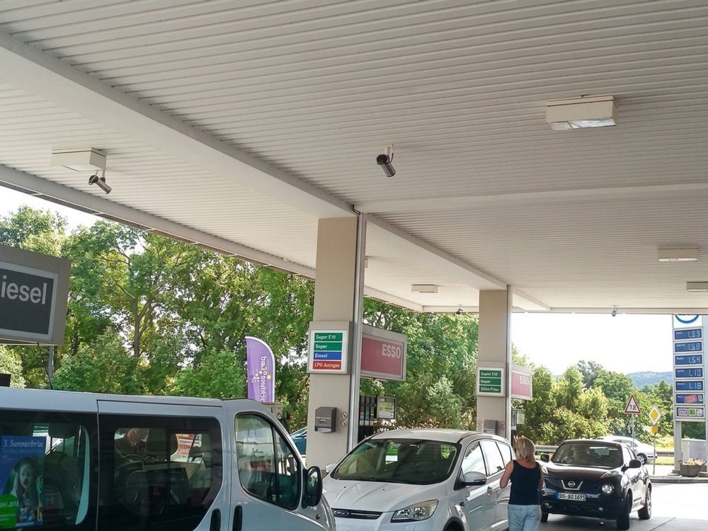 Tankstellenvorplatz mit Lieferwagen und Pkw unter einem Vordach, eine Person geht in der Nähe der Zapfsäulen entlang.