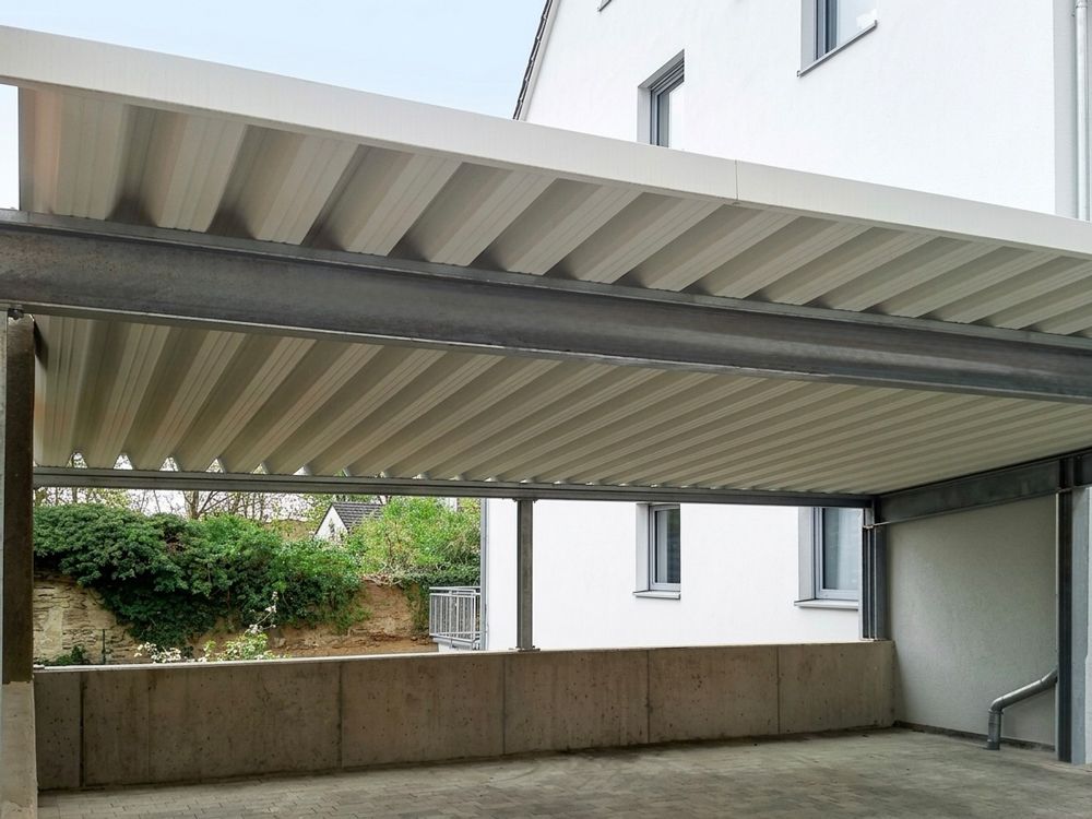 Überdachter Parkplatz mit Wellblechdach und Betonwänden neben einem weißen Gebäude.