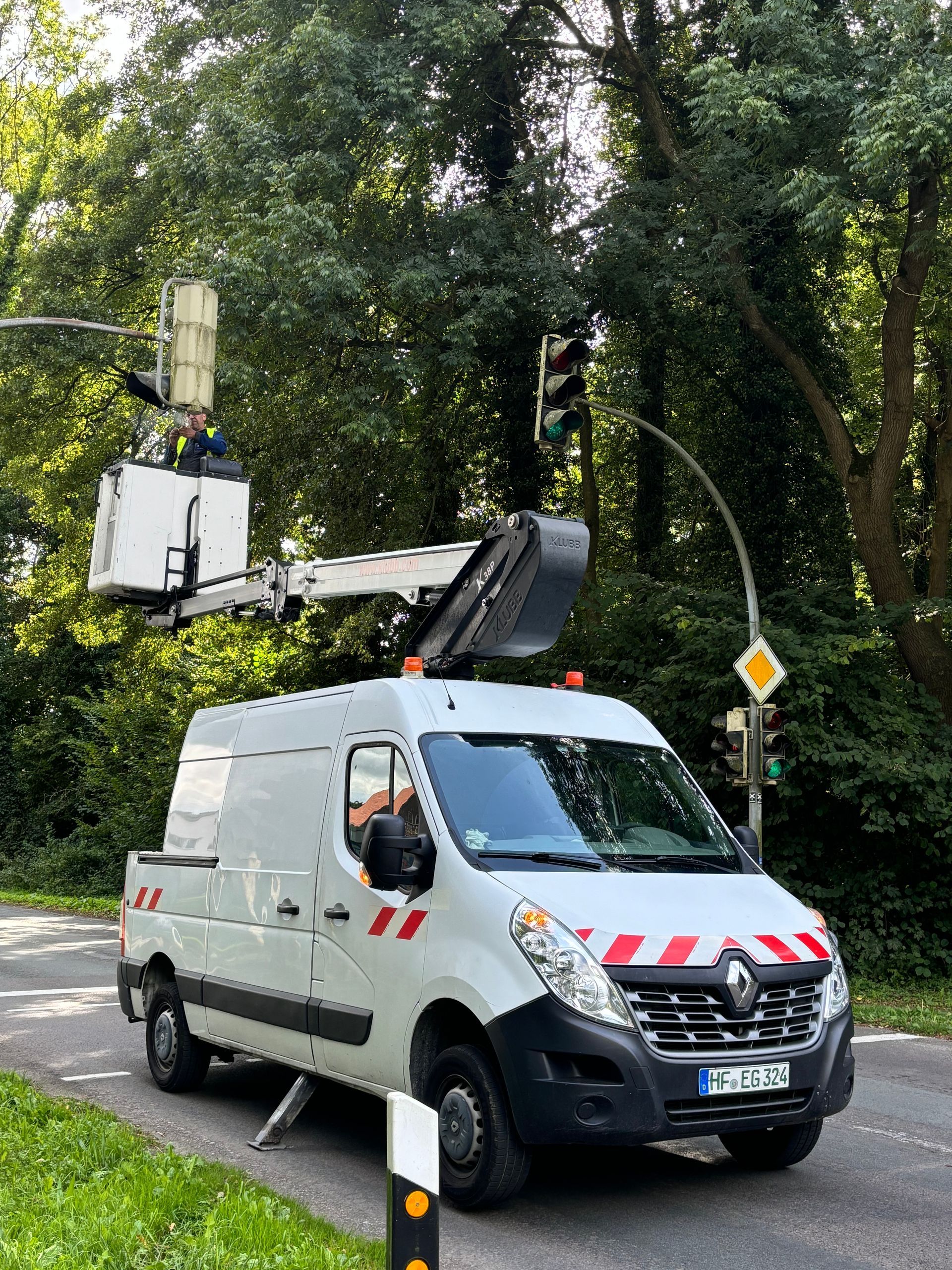 Ein Lieferwagen mit Hubarbeitsbühne steht neben einer von Bäumen gesäumten Straße; ein Arbeiter befindet sich im angehobenen Korb.