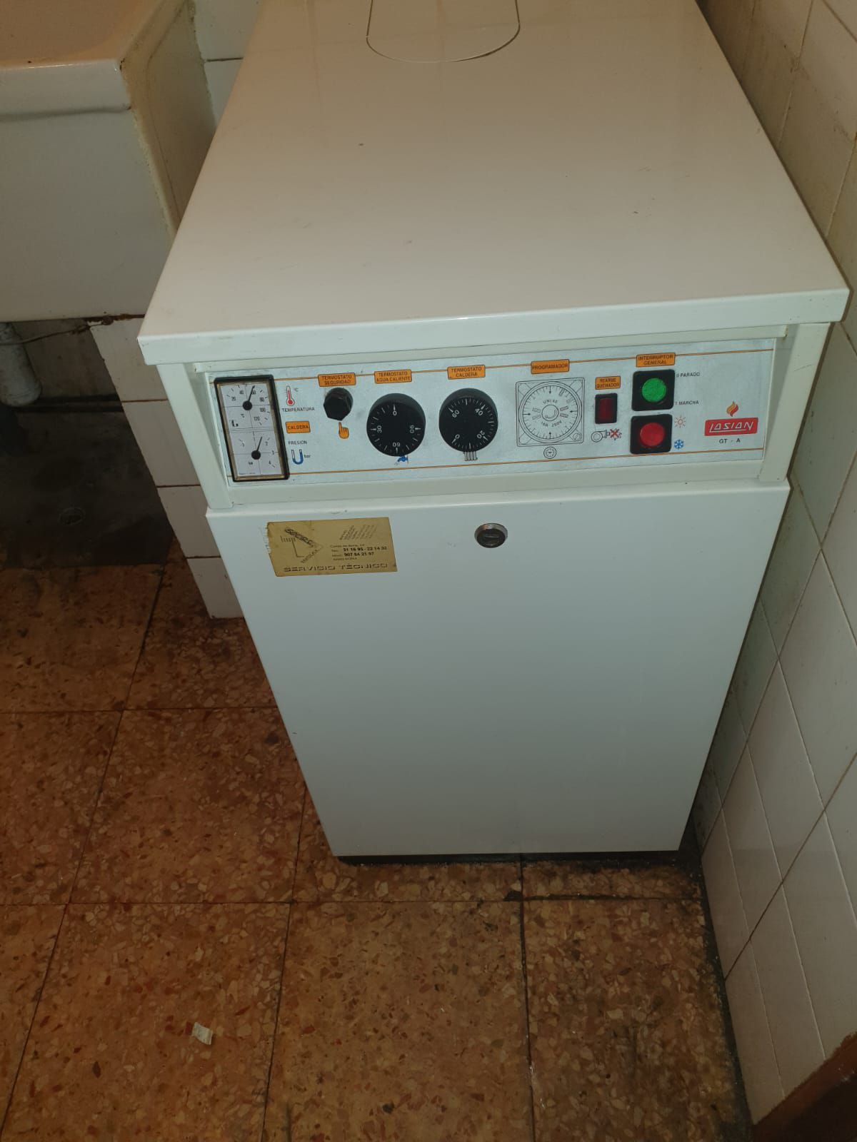 Calentador de agua blanco con diales y luces, colocado sobre un suelo de baldosas en una esquina del baño.