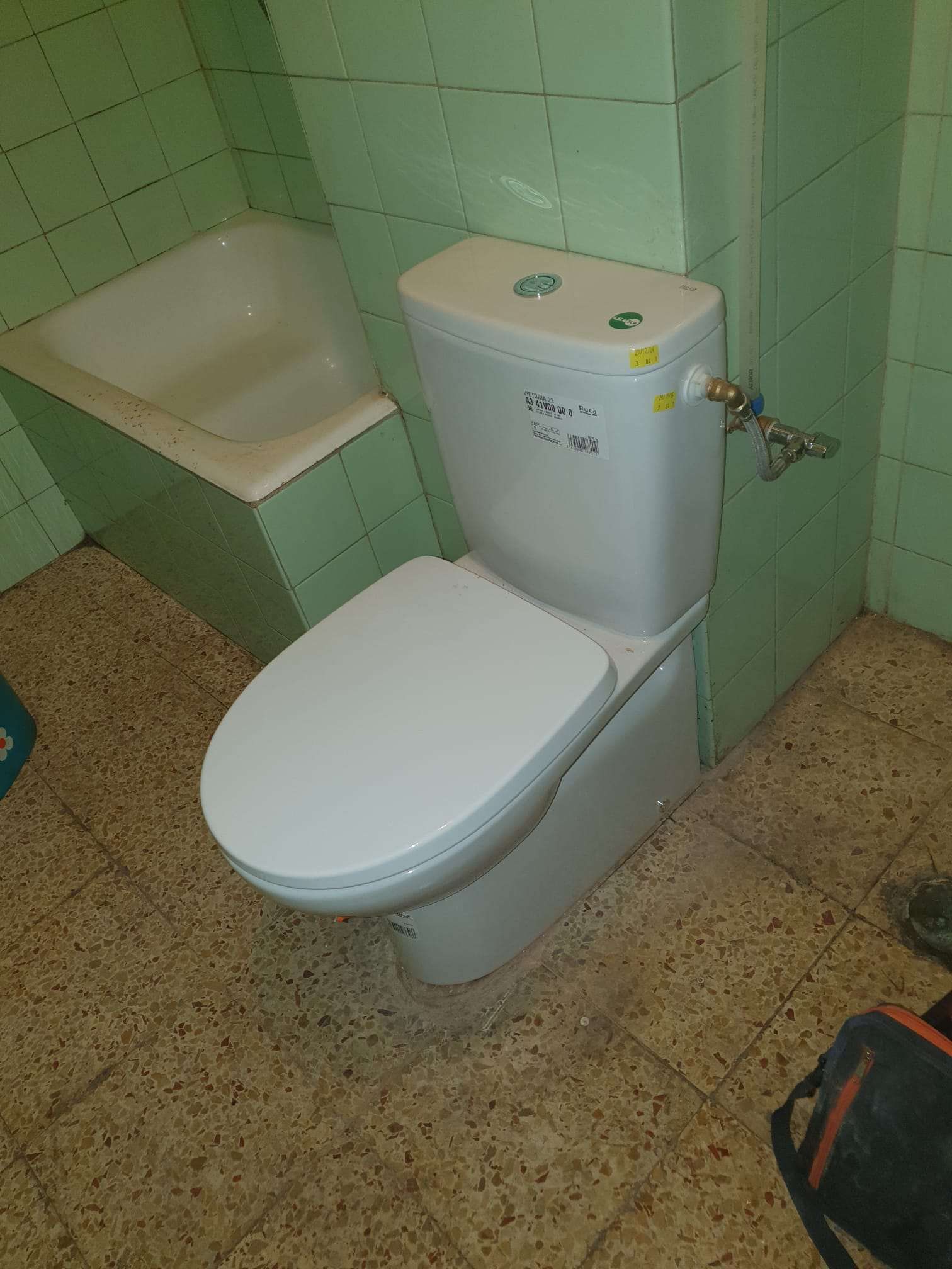 Inodoro con tapa y cisterna blancas en un baño con azulejos verdes; bañera al fondo.