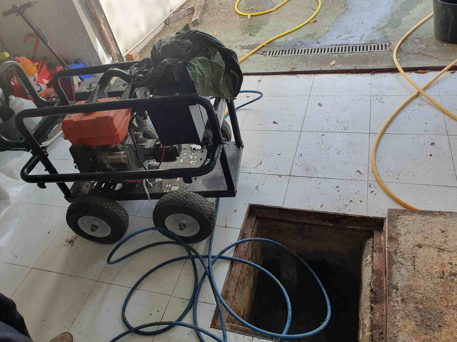 Una hidrolavadora sobre ruedas junto a una boca de alcantarilla abierta. Las mangueras están enrolladas sobre un suelo de baldosas.