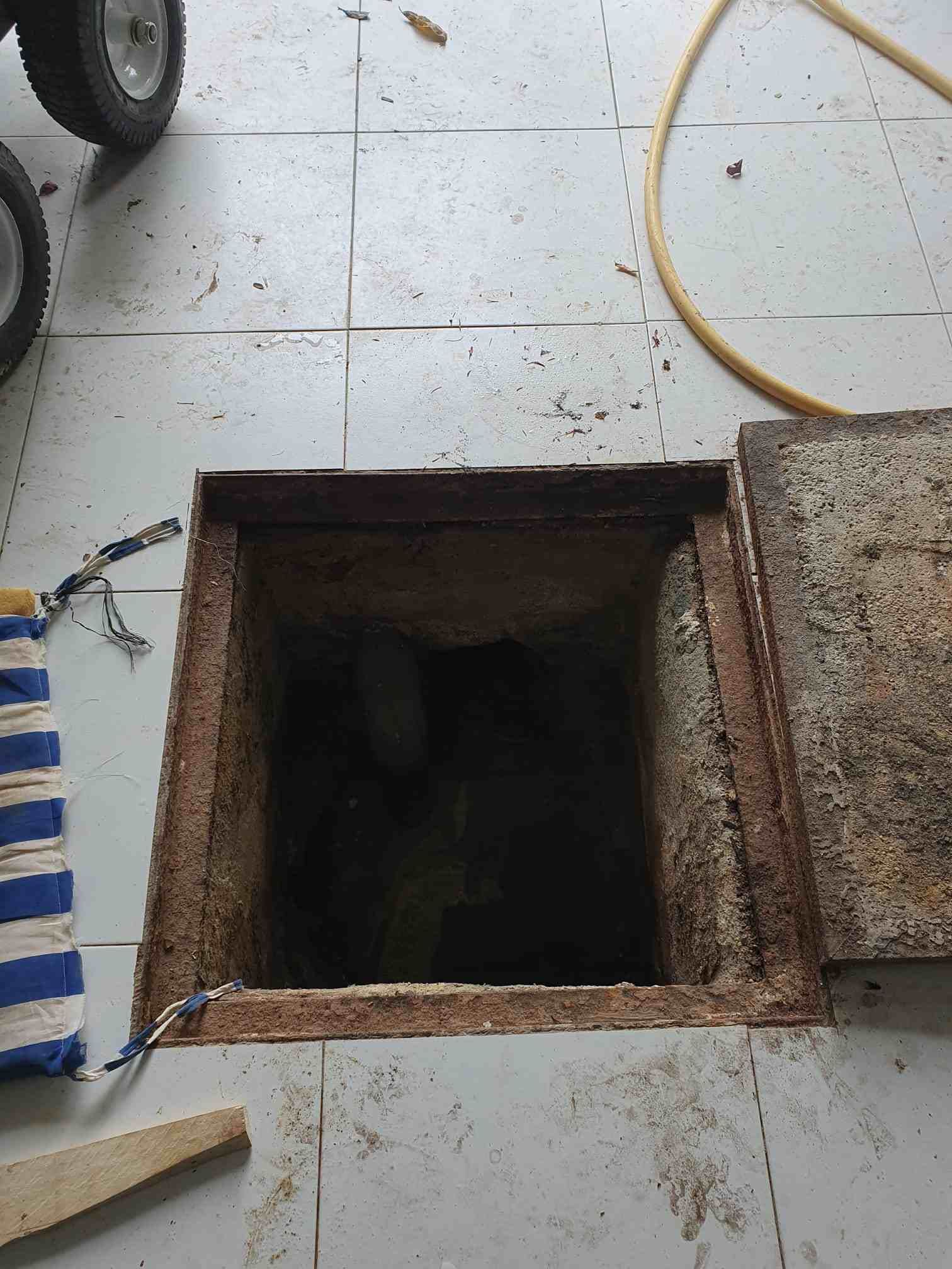 Agujero cuadrado abierto en el suelo de baldosas, parcialmente cubierto por una tapa metálica oxidada.