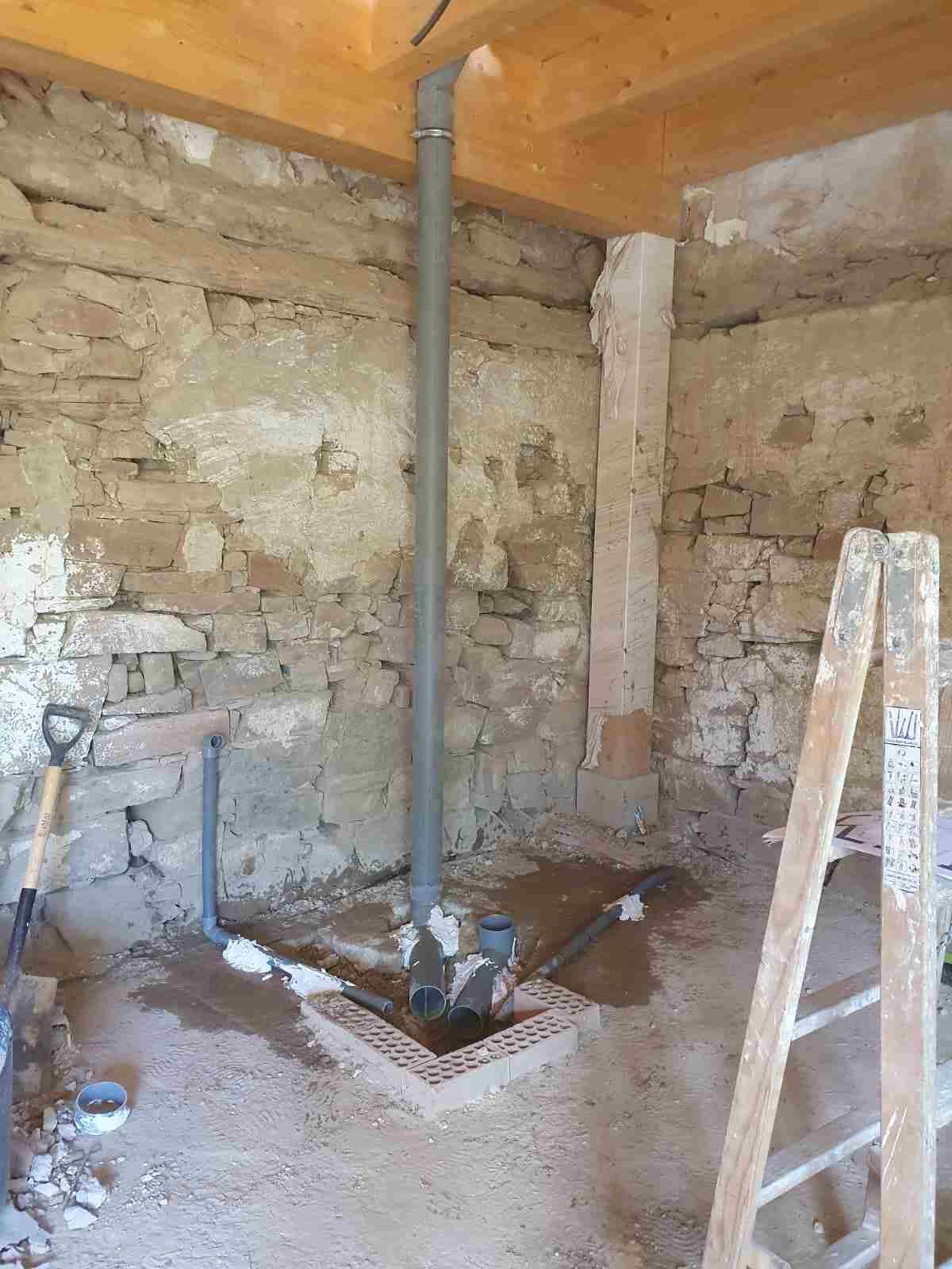 Trabajos de fontanería en una habitación con paredes de piedra; tubería gris vertical, rodeada de tuberías conectadas y una escalera.