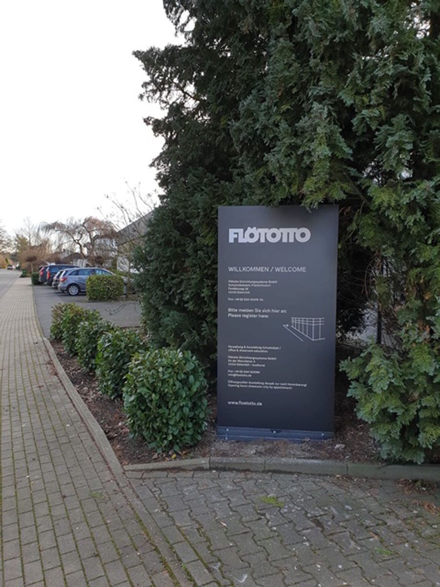 Am Rand einer Backsteinstraße neben einem Parkplatz steht ein schwarzes Schild.