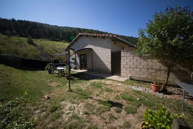 Edificio anexo rural de piedra con techo de tejas y puerta abierta, situado junto a un patio cubierto de hierba y una ladera.